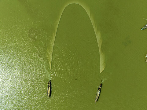Aerial view of two boats pulling a vast, arcing fishing net across the deep, verdant waters, Bogura, Rajshahi Division, Bangladesh.