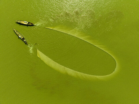 Aerial view of two slender boats navigating the vivid green waters around a pale, sandy oval formation, Bogura, Rajshahi Division, Bangladesh.