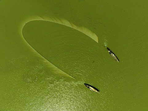Aerial view of two small boats navigating the serene emerald green waters, carving gentle ripples around a golden crescent of shallow land, Bogura, Rajshahi Division, Bangladesh.