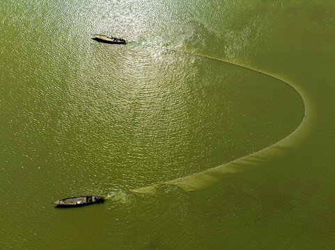 Aerial view of wooden boats casting a long, sinuous net through the vibrant green water under a shimmering sun, Bogura, Rajshahi Division, Bangladesh.