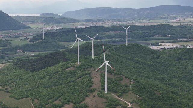 Aerial view of Monte Mesa Parco Eolico wind turbines standing on lush green hills overlooking the valley under an overcast sky in Rivoli Veronese, Veneto, Italy.