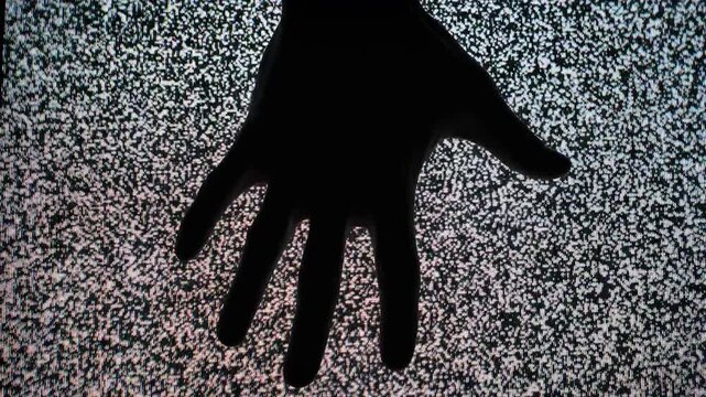 A dark silhouette of a human hand presses against a TV screen displaying chaotic heavy static noise. Concept of paranormal activity, retro horror atmosphere, and lost signal communication.