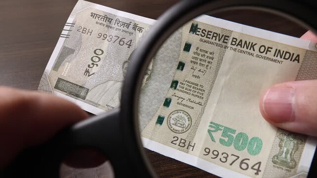 Examination of 500 Indian rupees (INR) cash money with magnfication glass, loupe. Concept of authenticity check, fight against counterfeiting of banknotes, anti fraud measures in India. Close up view.
