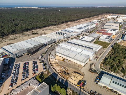 Aerial view of modern grey industrial warehouses contrasting with a dense green forest under a bright sky, Vagos, Aveiro, Portugal.