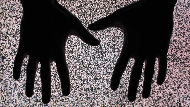 Silhouette hands slide across a flickering television screen displaying heavy static white noise. Concept of paranormal communication, horror atmosphere, and retro technology malfunction.