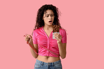 Shocked young African-American woman with sweet chocolate bar and measuring tape on pink background