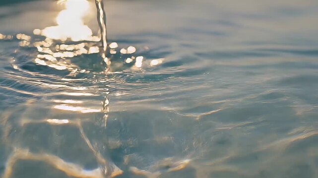 Water droplet impact creating ripples in clear blue surface with golden sunlight reflection