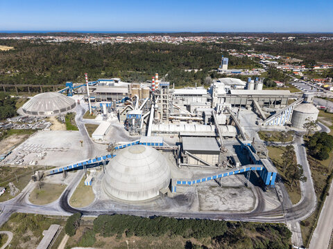 Aerial view of a vast cement factory, featuring prominent gray domes and towering stacks amidst lush green forests under a blue sky, Pataias, Leiria, Portugal.
