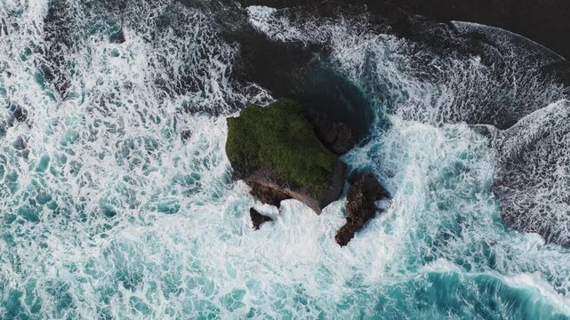 Aerial view of powerful ocean waves crashing against a green-topped rock formation at Mesra Beach in Kecamatan Tanjungsari, Daerah Istimewa Yogya, Indonesia.