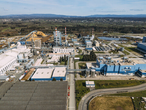 Aerial view of a large industrial plant with prominent blue structures, tall chimneys, and raw material piles, contrasting with the surrounding greenery, Cacia, Aveiro District, Portugal.