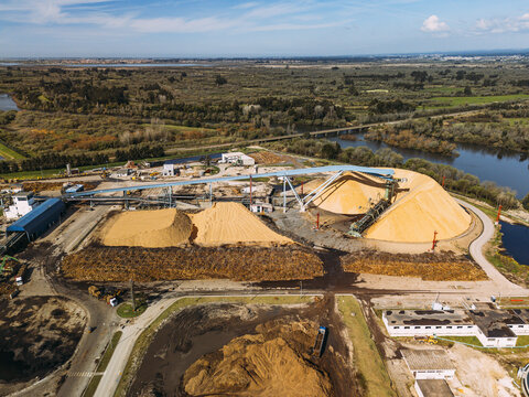 Aerial view of a wood processing facility, with golden timber piles and dark mulch contrasting a winding river and green forests, Cacia, Aveiro District, Portugal.