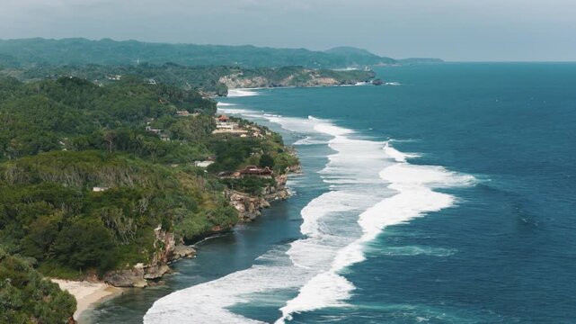 Aerial view of Mesra Beach featuring white waves crashing against the rugged rocky coastline and lush tropical green hills under a clear sky, Kecamatan Tanjungsari, Daerah Istimewa Yogya, Indonesia.