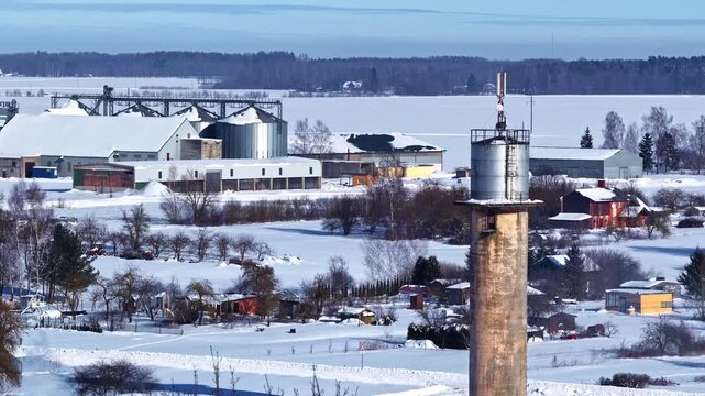 Aerial rising drone shot over snow-covered rural Latvian industrial town featuring Soviet-era chimney, grain silos, warehouses and vast frozen lake stretching to pine forest horizon