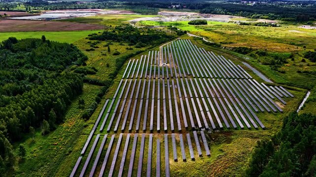 Aerial orbit drone shot circling large photovoltaic solar farm built on reclaimed land surrounded by lush green summer forest, meadows and wildflower fields in rural Latvia
