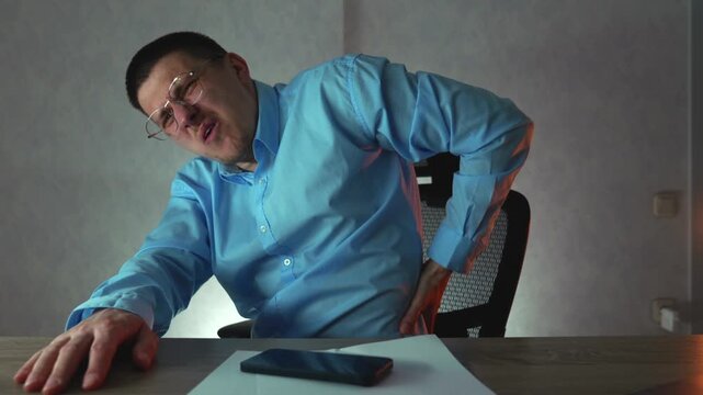 Tired office worker in dark office at evening holding his lower back in pain after prolonged sitting. Back pain, fatigue and unhealthy workplace habits concept.