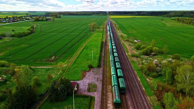 Aerial drone view of a long freight train crossing green farmland on a straight countryside railway, showing rural transport, spring fields, and expansive landscape.