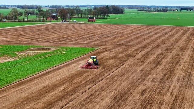 Aerial tracking green tractor with red seed drill performing spring seeding across vast brown plowed arable field surrounded by green crops and farmhouses in Latvia