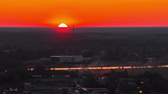 Aerial telephoto drone shot capturing half sun disk precisely on horizon over silhouetted Jelgava city with golden river reflection and fiery red-orange sky