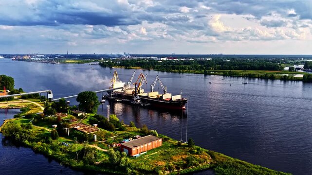 Cargo ship loading at riverside port with cranes and cityscape beyond