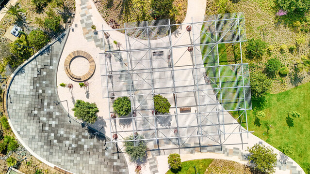 Urban park with abstract metal pergola casting geometric shadows over pathways, greenery, and modern seating, aerial view highlights contemporary landscape design and open space recreation