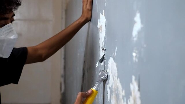 Asian painter man using scraper or spatula to remove old paint wall for cleaning, repair, preparing concrete wall before painting wall at home. Scraping to remove paint plaster peel. Painting concept.