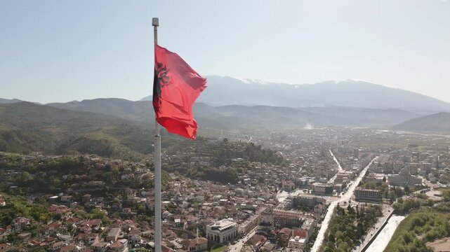 Aerial static view Albanian national flag prominently wave atop stone fortification at Berat Castle - Kalaja e Beratit under clear blue sky, overlooking lush, forested hillside and Berat cityscape