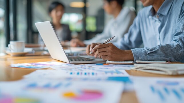 Focused Business Dynamics: A detailed view of a group engaged in a project discussion, the main focal point being a person meticulously working on a laptop surrounded by analytical charts.