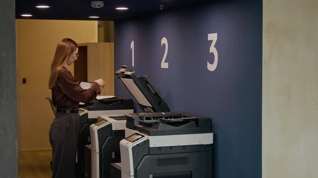 Young female office worker holding folder, walking to printing station and copying business document with modern multifunction printer