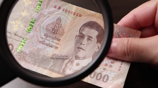 Examination of 1000 Thai baht (THB) cash money with magnfication glass, loupe. Concept of authenticity check, fight against counterfeiting of banknotes, anti fraud measures in Thailand. Close up view.
