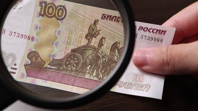 Examination of 100 Russian ruble (RUB) cash money with magnfication glass, loupe. Concept of authenticity check, fight against counterfeiting of banknotes, anti fraud measures in Russia. Close up view