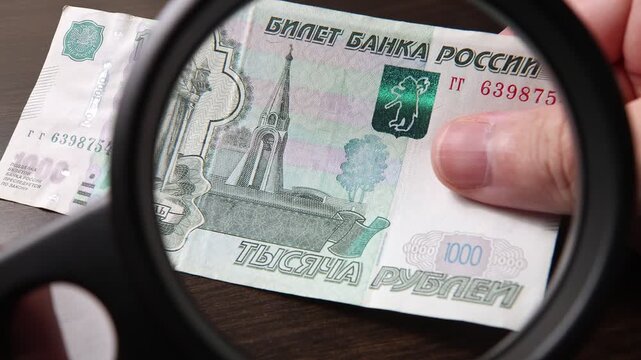 Examination of 1000 Russian ruble cash money with magnfication glass, loupe. Concept of authenticity check, fight against counterfeiting of banknotes, anti fraud measures in Russia. Close up view.