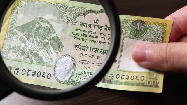 Examination of 100 Nepalese rupee (NPR) cash money with magnfication glass, loupe. Concept of authenticity check, fight against counterfeiting of banknotes, anti fraud measures in Nepal. Close up view