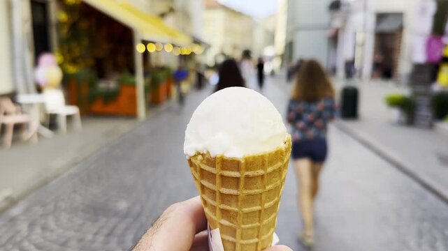 POV of male hand holding an ice cream cone while walking along city street. Blurred pedestrians pass by in the background, creating a lively urban atmosphere and emphasizing the treat in arm