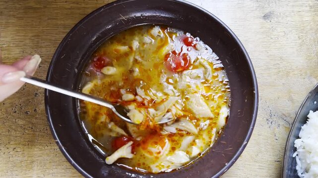 Top view of female hand gently stirs tom yum soup with spoon at restaurant table. Steam rises from the aromatic broth as the ingredients swirl together capturing a cozy dining moment. Slow motion
