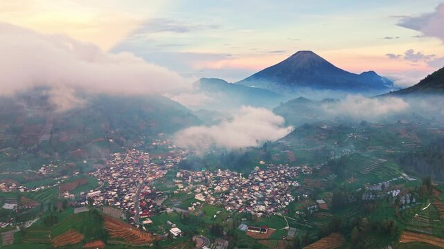 Mountain village nestled in misty valley. Clouds drift around lush green slopes