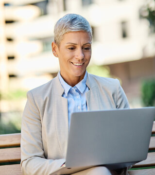 Portrait of a young businesswoman woman using a laptop computer and sitting on a bench in a coty park, surrounded by modern corporate office buildings