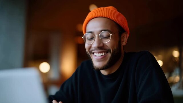 Smiling young man working on laptop in modern cozy cafe interior, freelancer lifestyle concept, casual creative professional with glasses and orange beanie, remote work