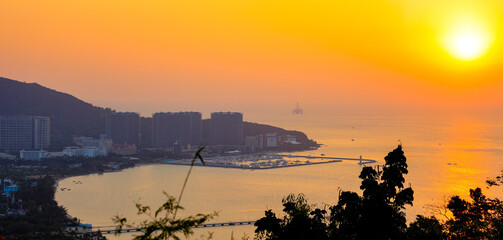 aerial view of golden sunset illuminating a coastal bay, where modern high-rise buildings and a yacht-filled marina sit nestled between hills and the shimmering sea, background image © Echo