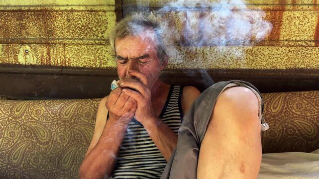 Portrait of homeless adult man sitting on worn couch placed and smoking outdoor. The setting underscores neglect and impermanence, presenting a raw and unfiltered glimpse of difficult living situation