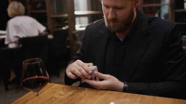 POV handheld shot of bearded young Caucasian man wearing suit presenting diamond engagement ring in jewelry box across table, proposing marriage in luxury restaurant setting