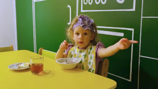 Moskow, Russia, 21.08.2025 Caucasian girl sitting at yellow table eating lunch. Toddler wearing bib pointing at something during meal. Child in kitchen having healthy food and drink time.