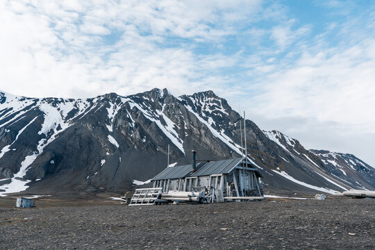 View of the historic Bamsebu wooden hut standing on a rocky plain against a backdrop of snow-capped mountains in Svalbard, Svalbard and Jan Mayen.