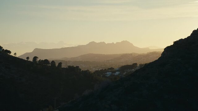 Golden evening light settles over the mountain valley above Moraira, Spain, with hazy ridgelines, hillside homes, and a peaceful Mediterranean atmosphere ideal for travel content.