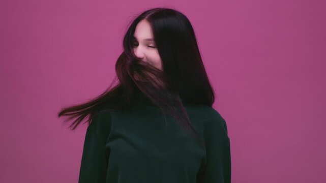 Beautiful cheerful brunette young girl wearing green pink sweater posing isolated on pink wall. Dancing shaking head with flowing hair. Teenage woman shakes her straight black long hair, slow motion.