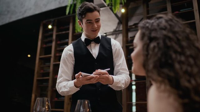 Medium slowmo shot of courteous young Caucasian waiter in classic uniform with bowtie taking order on notepad from unrecognizable female guest sitting at table in fine dining restaurant