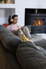 Obraz premium Non-binary person sitting on gray sectional sofa wearing headphones and using tablet by fireplace