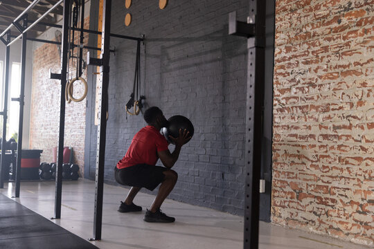 African American man squatting while holding black medicine ball wearing red shirt in gym
