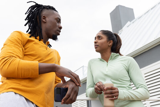 Diverse friends standing on rooftop balcony leaning on rail holding phone and bottle in sports tops