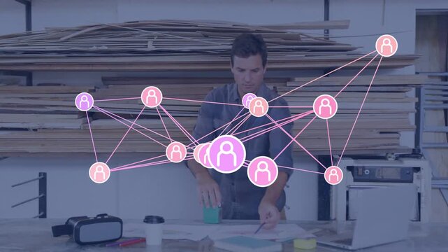 Mature man in denim shirt working at wood shop bench arranging plans under pink network overlay