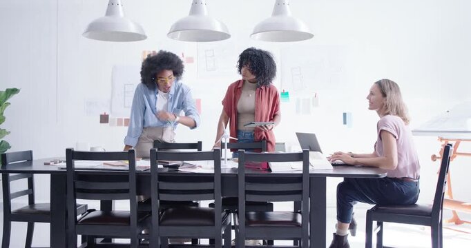 Diverse female team reviewing layouts at table, moving swatches to wall, choosing palette on laptop